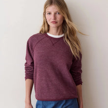 Load image into Gallery viewer, A woman modeling a burgundy and maroon horizontally striped long-sleeve crewneck sweater over a white t-shirt, paired with a blue denim skirt and holding an olive green jacket, set against a plain off-white wall.
