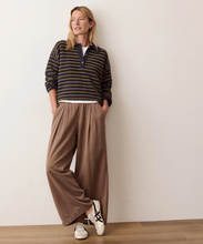 Load image into Gallery viewer, A woman stands against a plain off-white wall wearing a dark striped long-sleeve henley top, wide-leg brown pants, and white sneakers.
