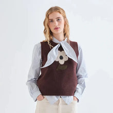 Load image into Gallery viewer, A woman modeling a dark brown knit sweater vest with a white bow and a white flower applique on the chest, worn over a light blue collared shirt and cream trousers, against a plain white studio background.
